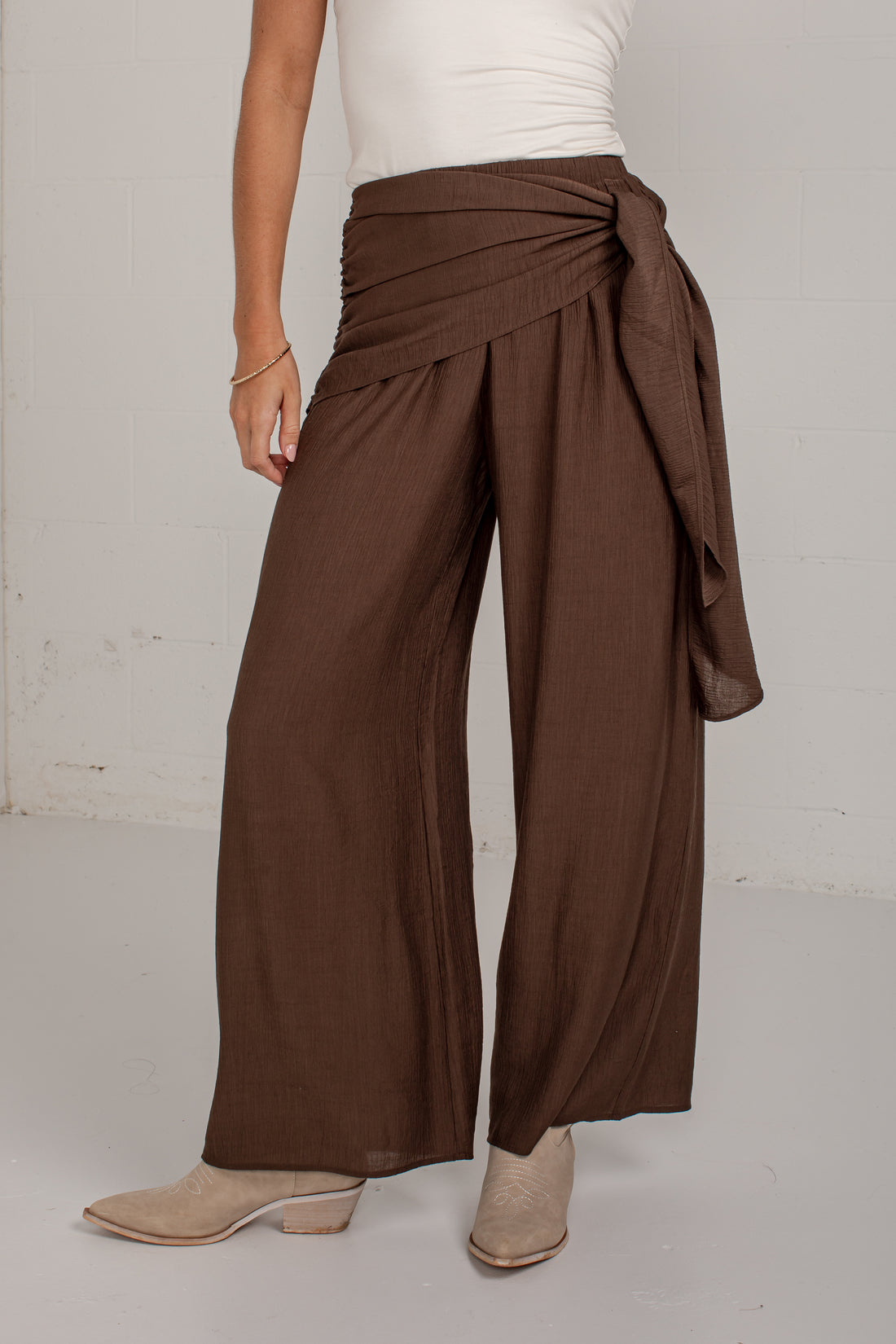 A person wearing wide-leg draped pants in a medium brown color, styled with a white top and beige ankle boots, standing against a light gray wall.