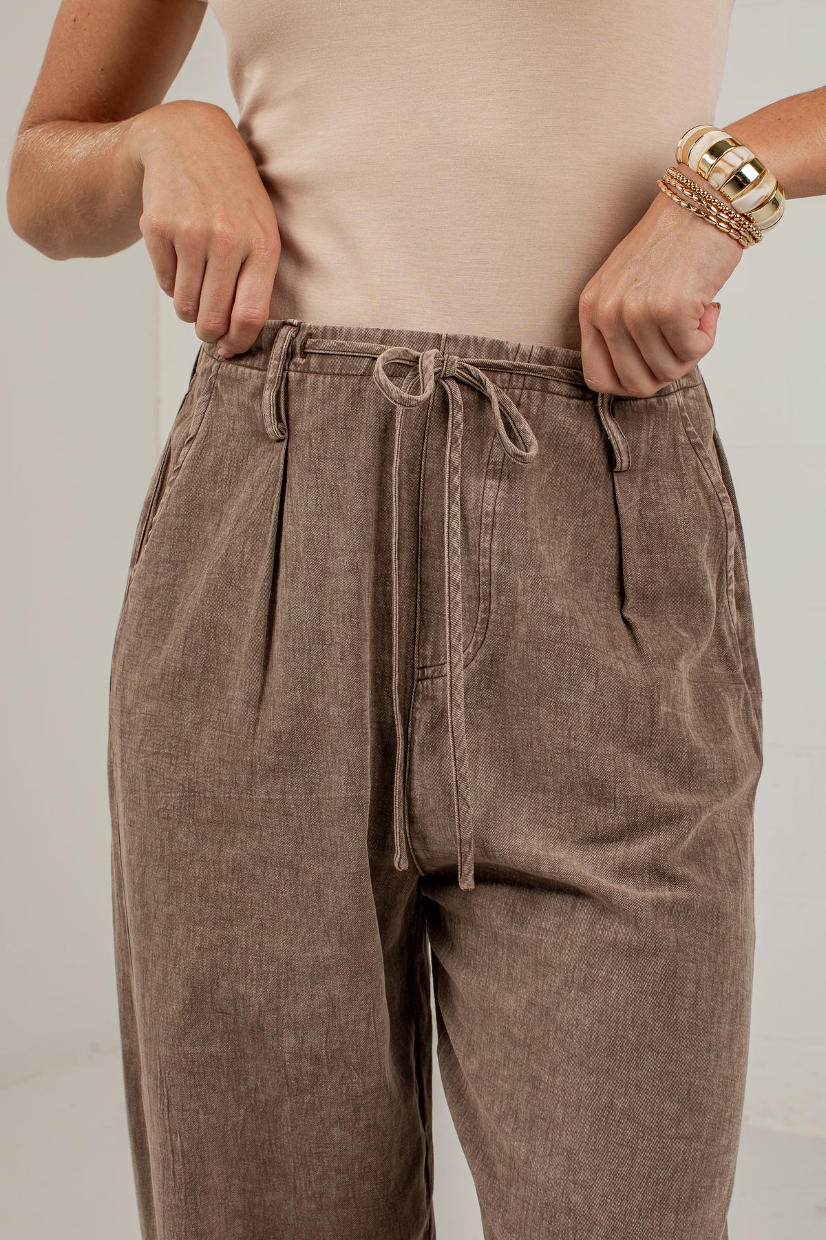A close-up of a person wearing high-waisted taupe corduroy trousers with a drawstring waistband, paired with a beige top and gold bracelets.
