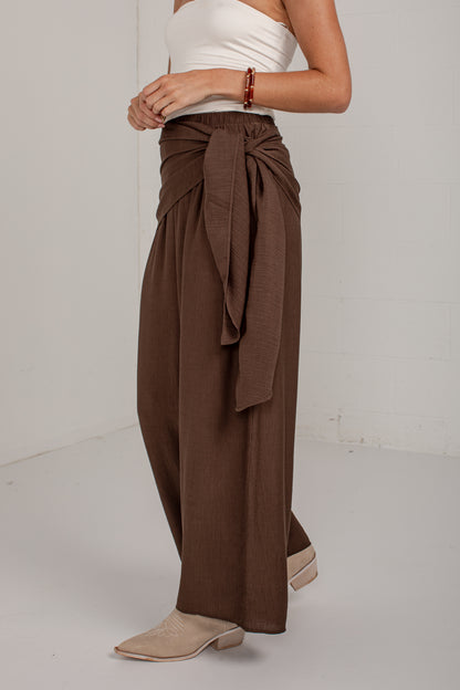 A model wearing high-waisted, wide-leg brown pants with a large side tie, styled with a white strapless top and beige pointed-toe mules against a white studio background.