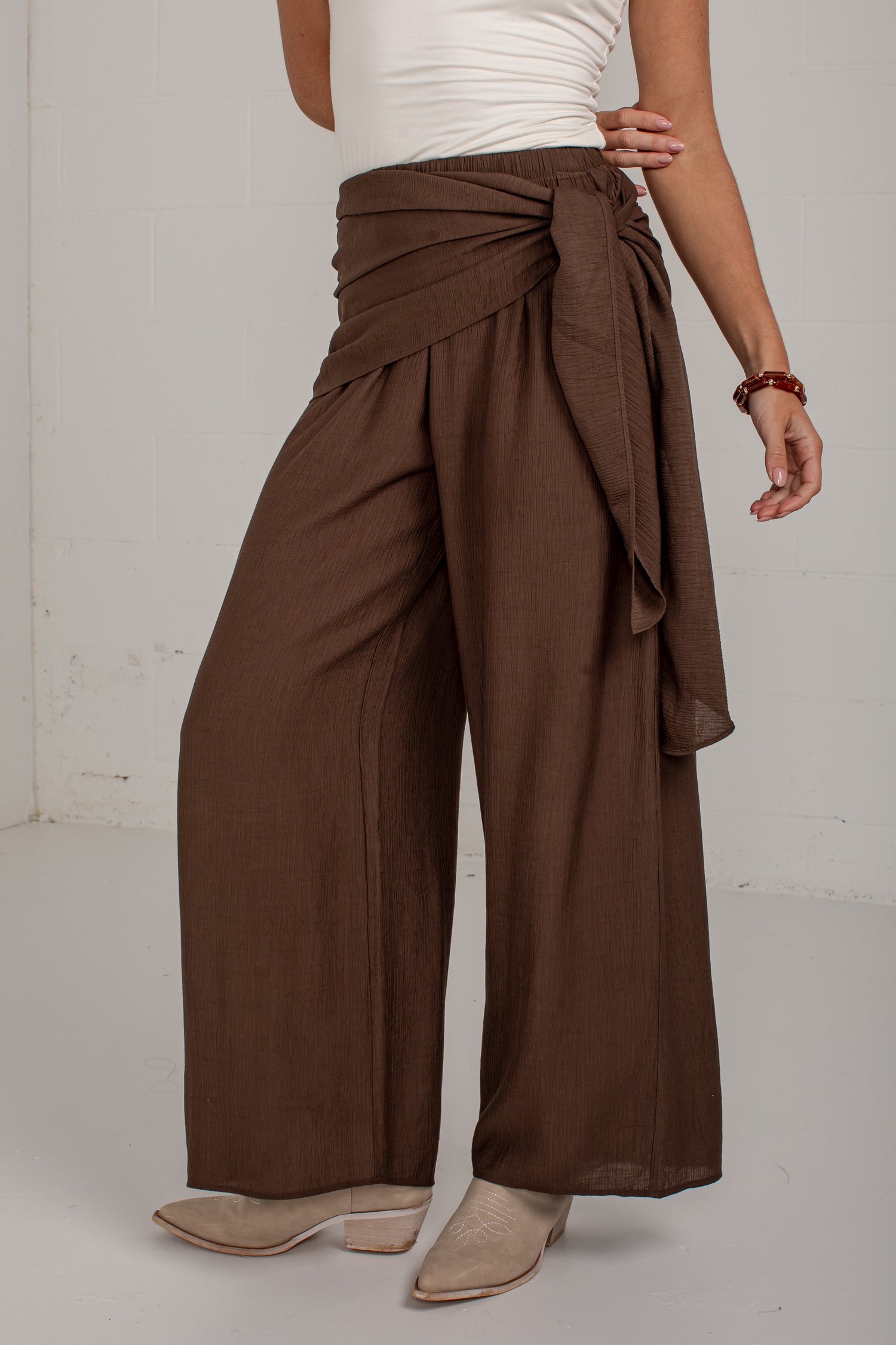 A model wearing high-waisted, wide-leg brown pants with a draped wrap waist and tie detail, styled with a white top and beige heeled mules against a white studio background.