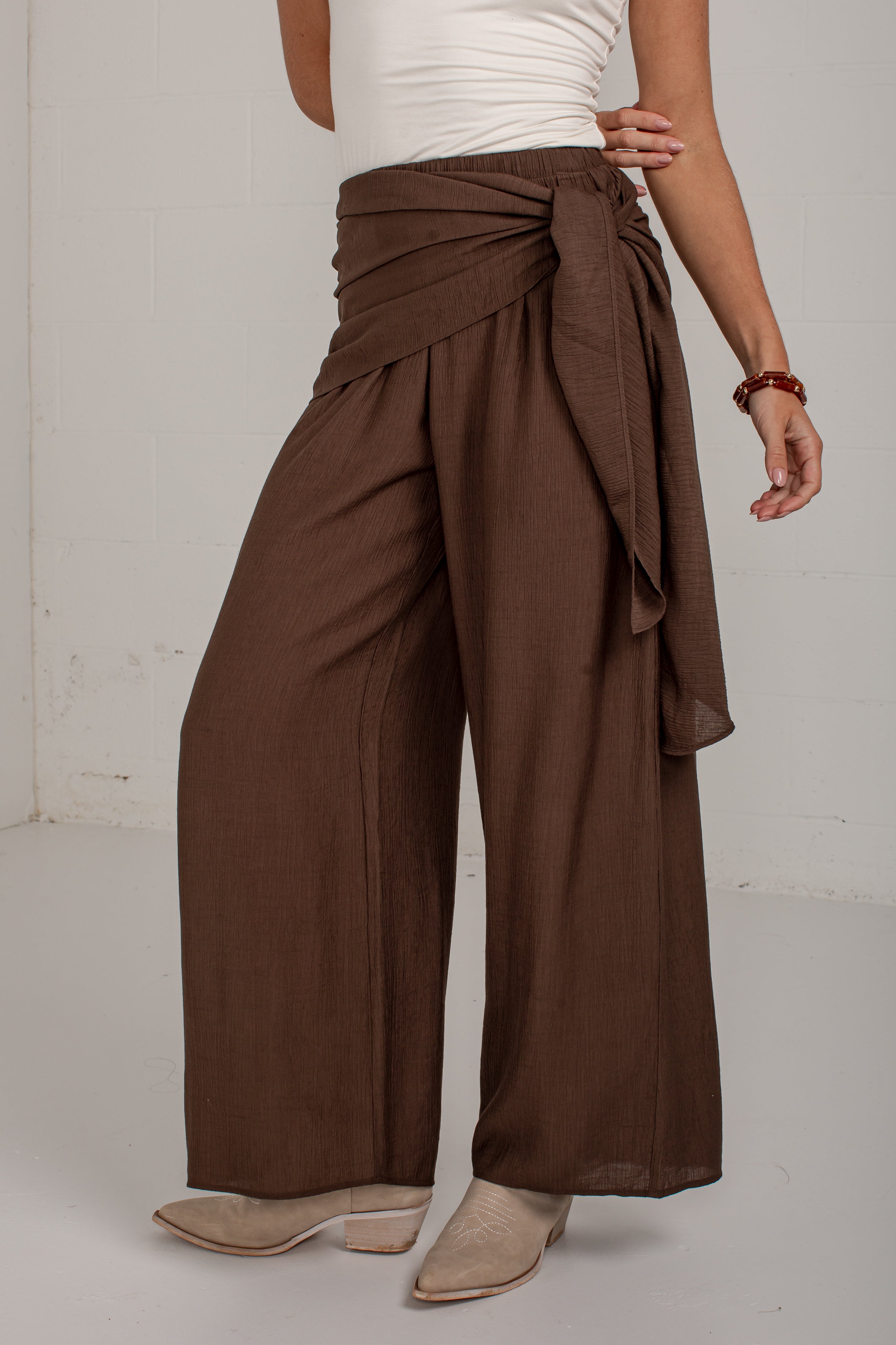 A model wearing high-waisted, wide-leg brown pants with a draped wrap waist and tie detail, styled with a white top and beige heeled mules against a white studio background.