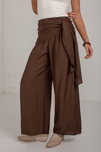 A model wearing high-waisted, wide-leg brown pants with a draped wrap waist and tie detail, styled with a white top and beige heeled mules against a white studio background.