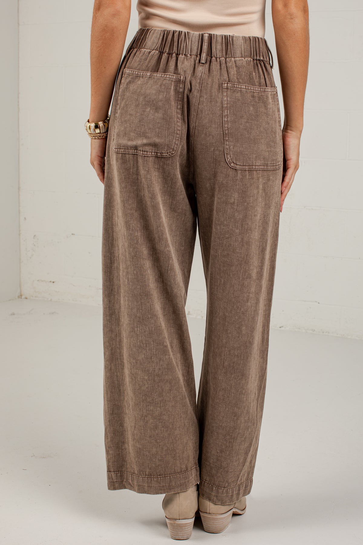 A person shown from the back wearing wide-leg brown pants with an elastic waistband and large patch pockets, photographed in a plain indoor studio setting.
