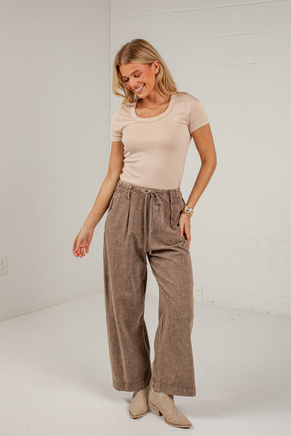 A woman modeling a pair of high-waisted, wide-leg taupe corduroy pants with a drawstring waist, paired with a beige t-shirt and beige ankle boots, standing against a white brick wall.