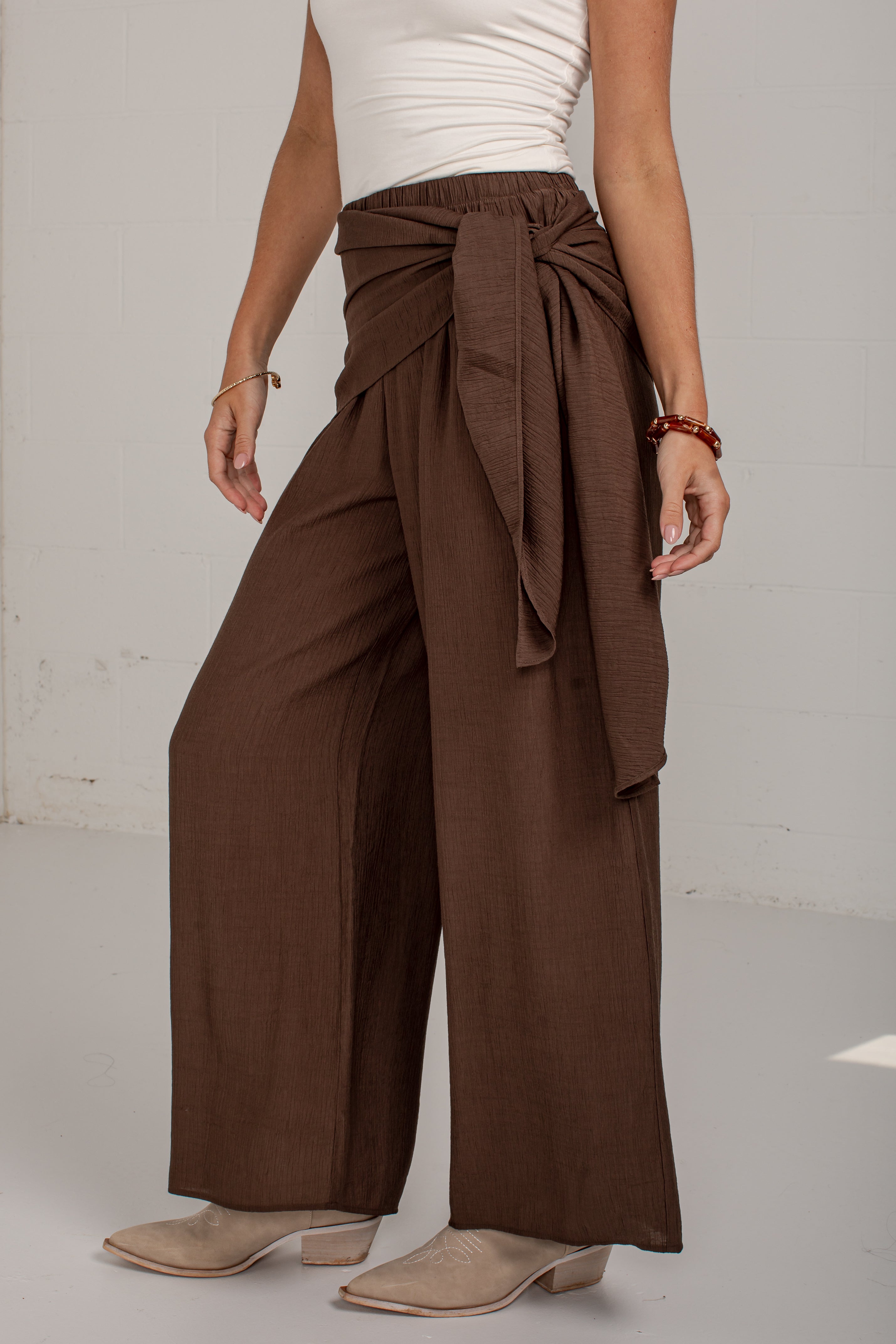 A model wearing high-waisted, wide-leg brown pants with a matching fabric tie belt at the waist, styled with a white ribbed tank top and beige heeled mules.