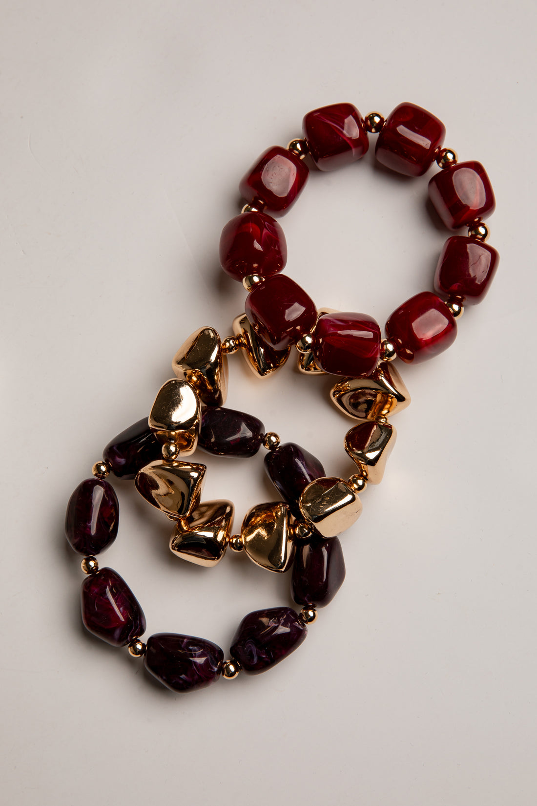 A multi-strand beaded necklace featuring deep red and dark purple marbled beads, accented with shiny gold metallic heart-shaped charms, displayed on a light gray background.
