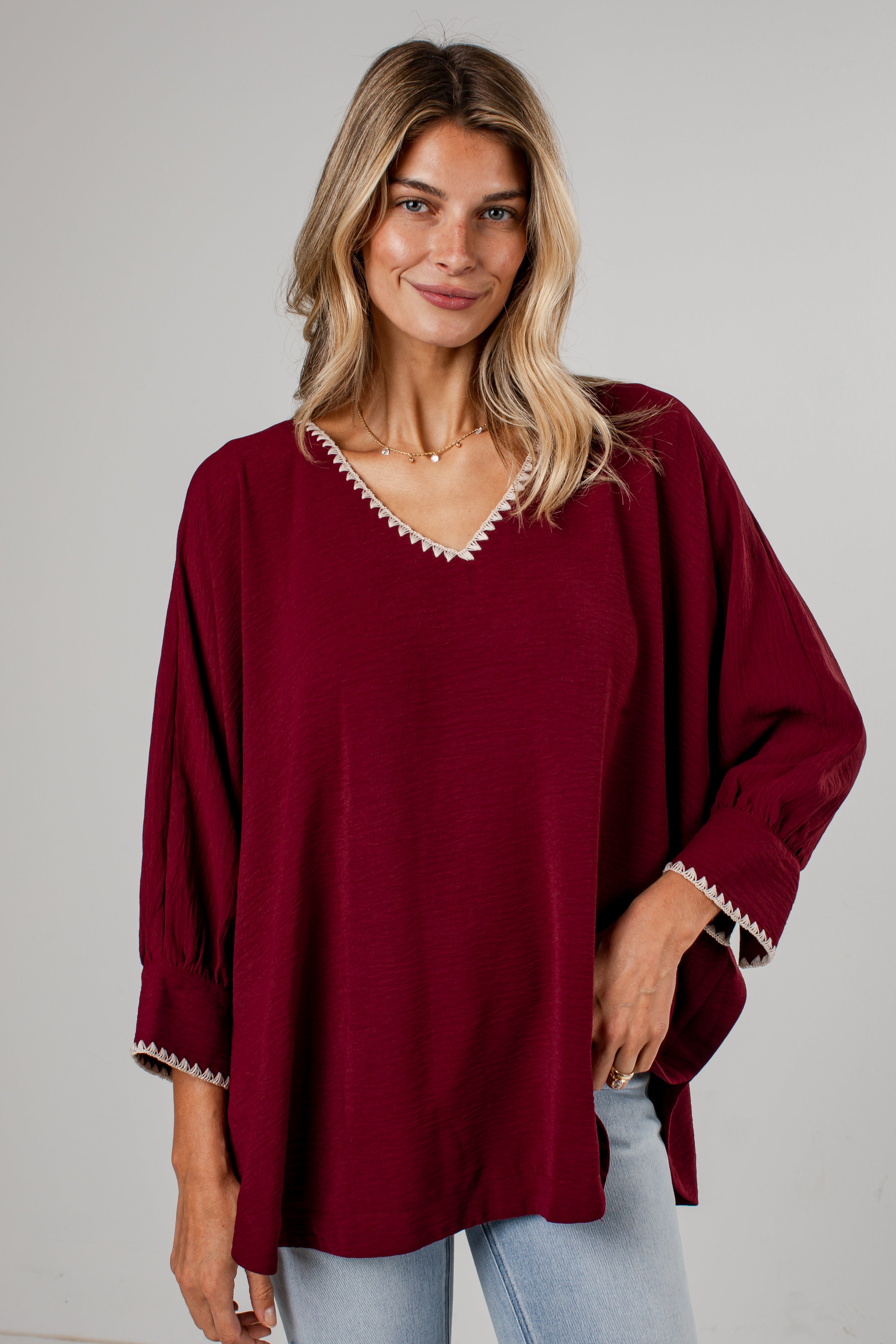 A woman wearing a loose-fitting burgundy blouse with white embroidered trim along the V-neck and cuffs, paired with light-wash blue jeans, standing against a light gray wall.