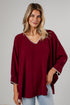 A woman wearing a loose-fitting burgundy blouse with white embroidered trim along the V-neck and cuffs, paired with light-wash blue jeans, standing against a light gray wall.