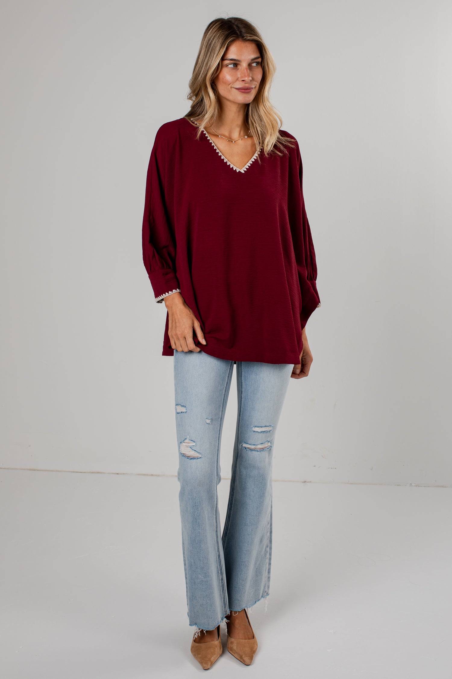 A woman modeling a loose-fitting burgundy long-sleeve top with a decorative white beaded trim along the V-neckline and cuffs, paired with light-wash distressed flare jeans and tan pointed-toe flats, against a plain light gray studio background.