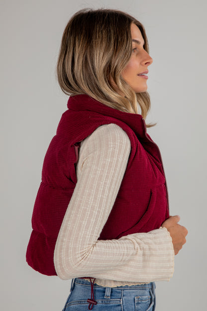 Woman wearing a red puffer vest over a beige sweater on a plain background