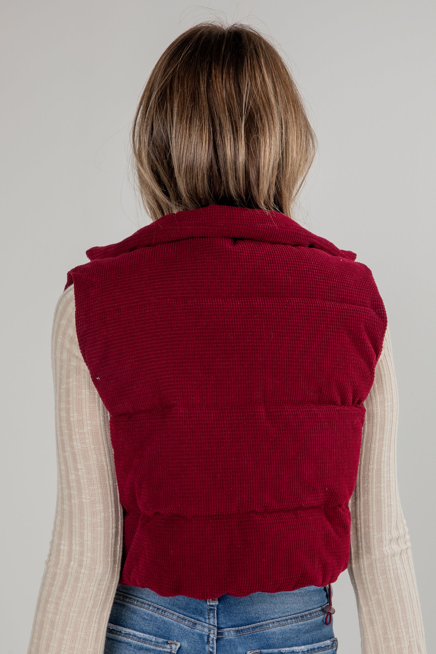 Person wearing a red puffer vest over a beige sweater and blue jeans on a plain background