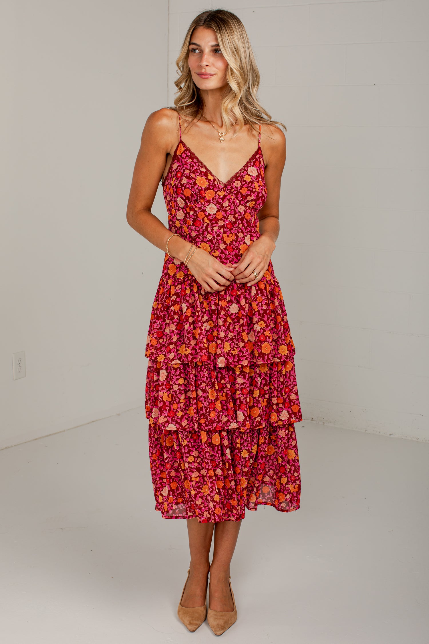 A woman modeling a tiered midi dress with a vibrant red, pink, and orange floral pattern, standing against a plain white studio background.