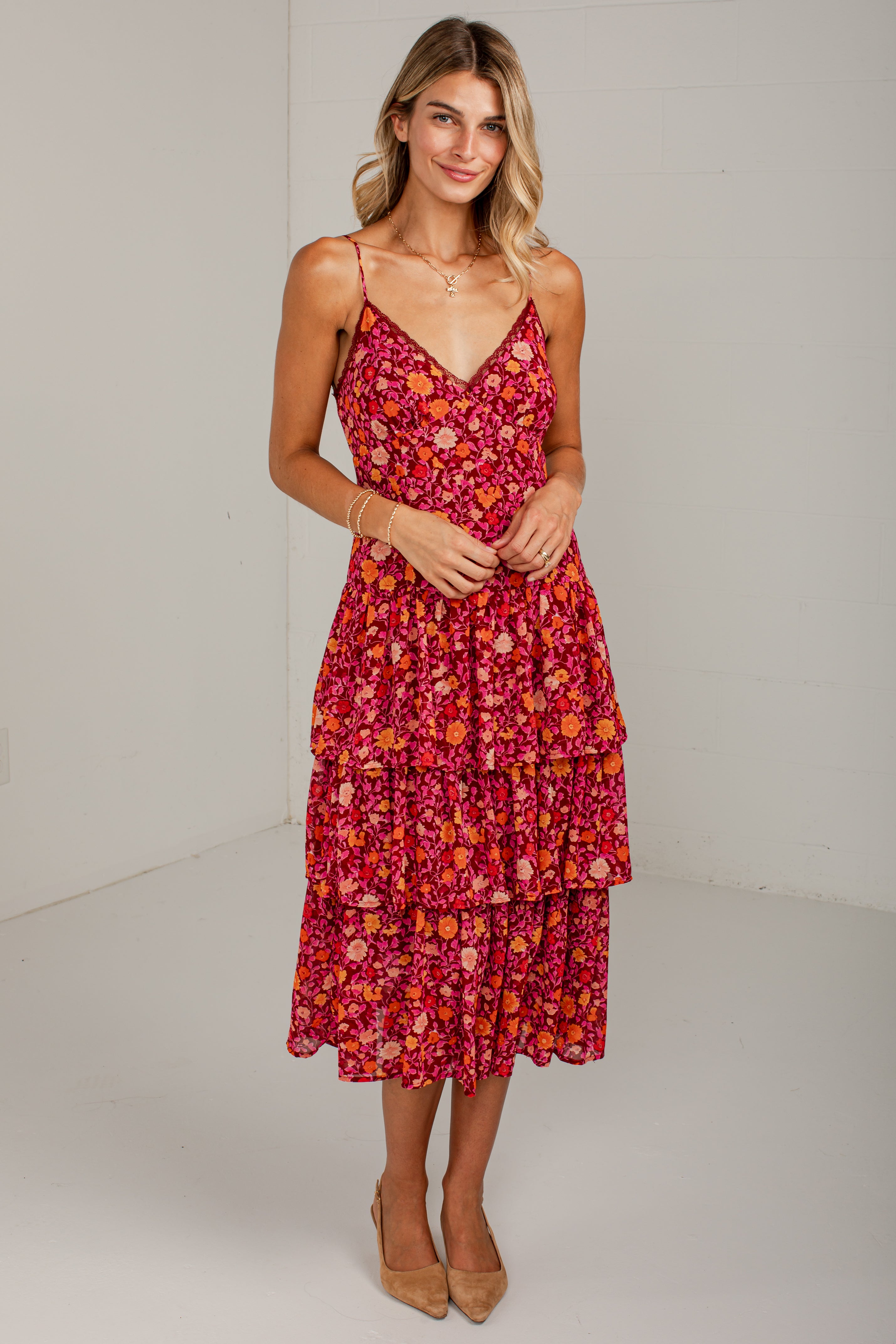 A woman modeling a tiered midi dress with a vibrant red, orange, and pink floral pattern, featuring spaghetti straps and a V-neckline, set against a plain white studio background.