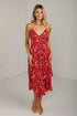 A woman modeling a red floral spaghetti-strap midi dress with a tiered ruffle hem, paired with beige pointed-toe heels, standing against a white studio wall.