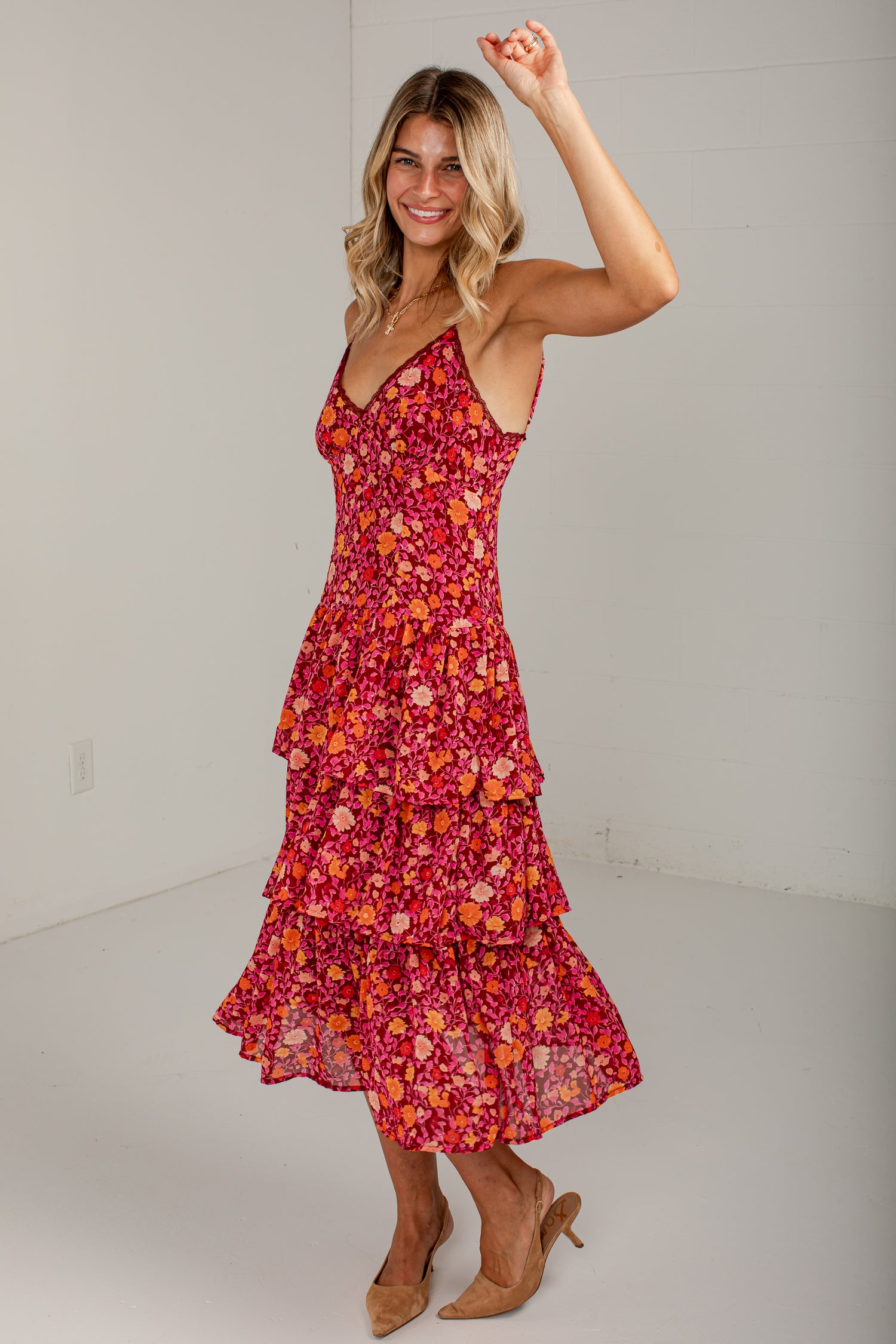 A woman modeling a sleeveless tiered midi dress with a vibrant red, orange, and pink floral print, paired with tan high heels in a bright studio setting.