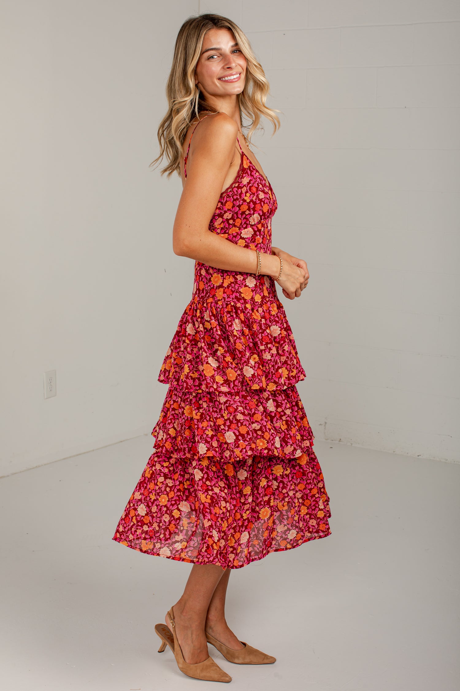 A woman modeling a sleeveless, tiered midi dress with a vibrant red, orange, and pink floral pattern, paired with tan heels, set against a plain white studio background.