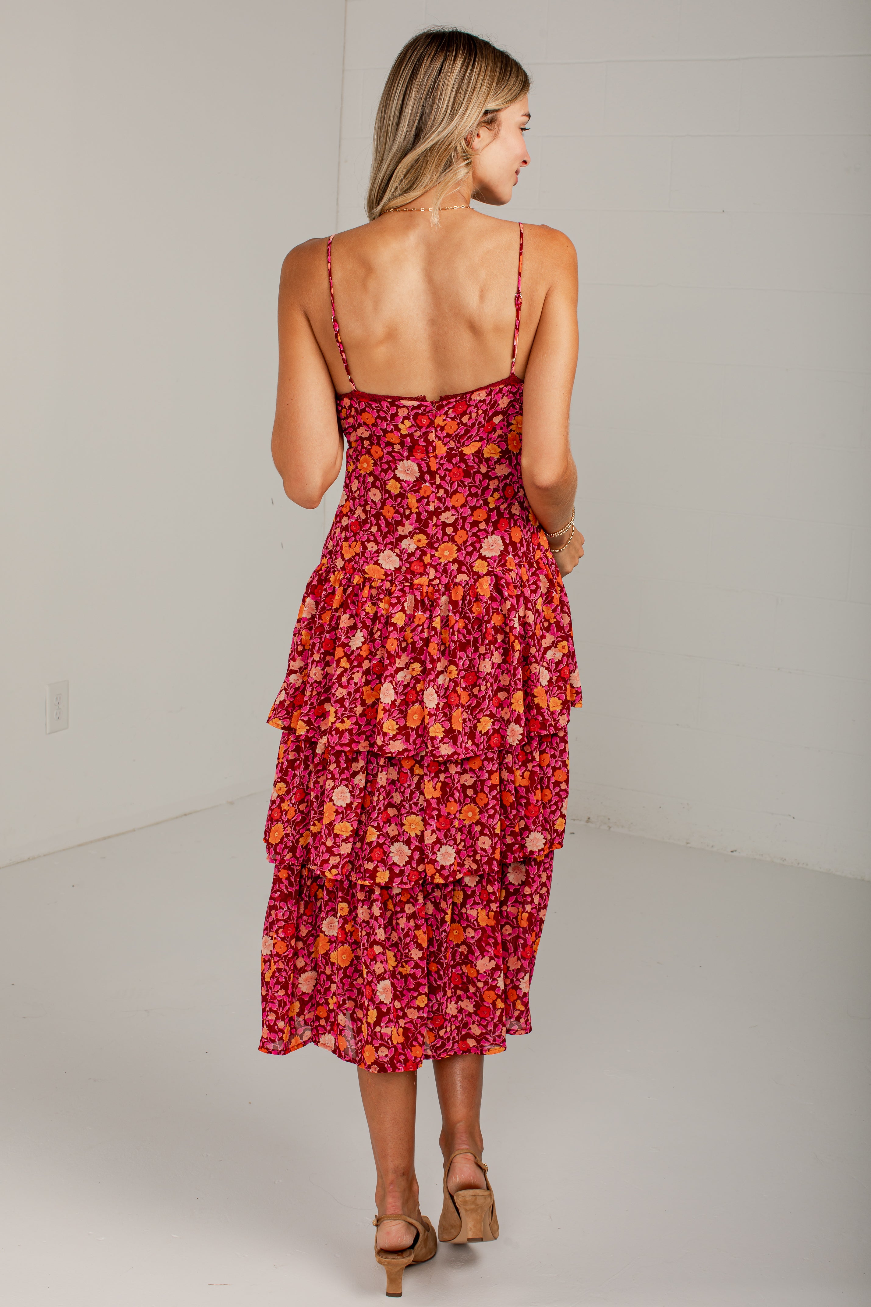 A woman modeling a tiered midi dress with a vibrant red, pink, and orange floral pattern, shown from the back in a studio setting.