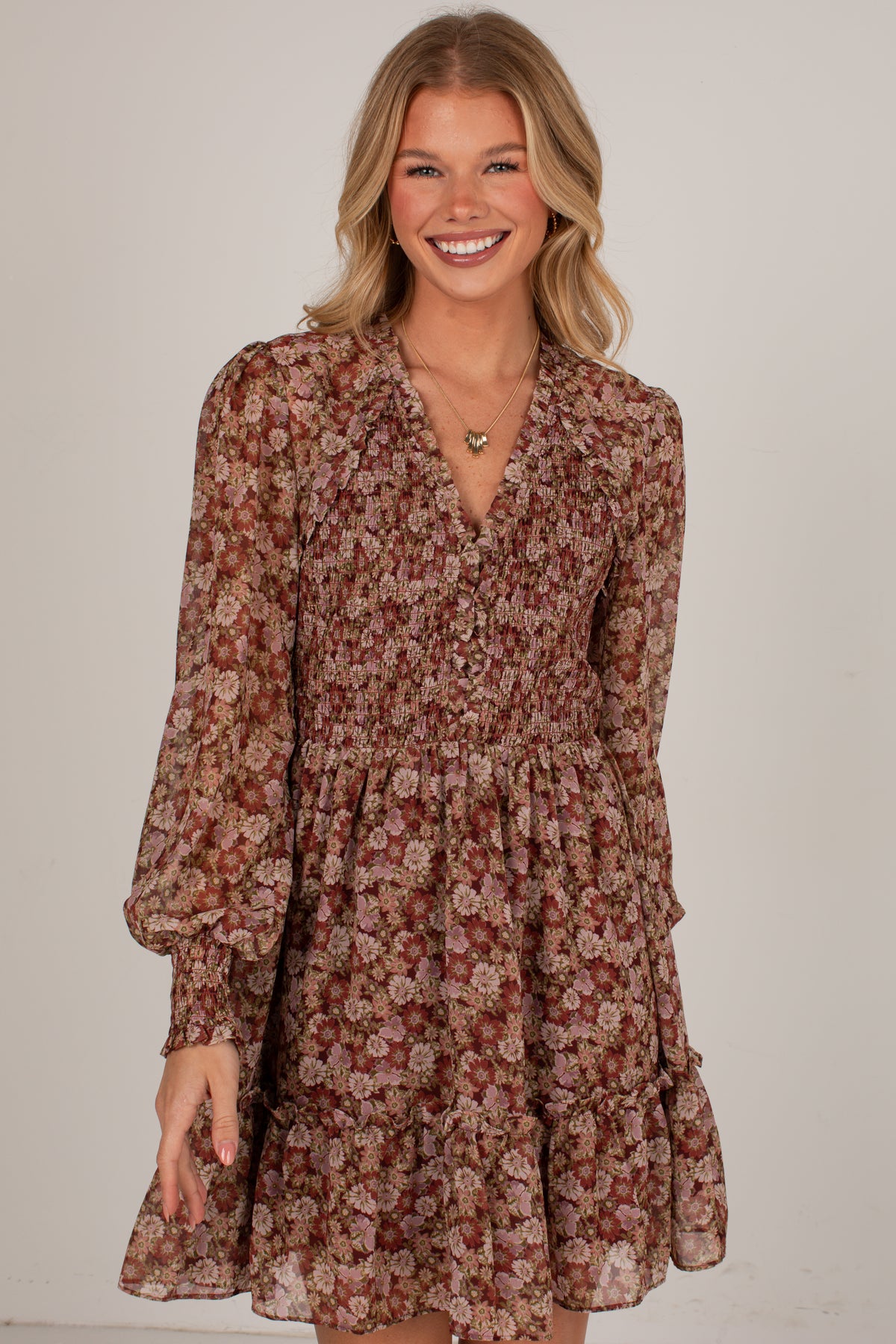 A smiling woman wearing a long-sleeve floral mini dress with a smocked bodice and tiered skirt, photographed against a plain light gray studio background.