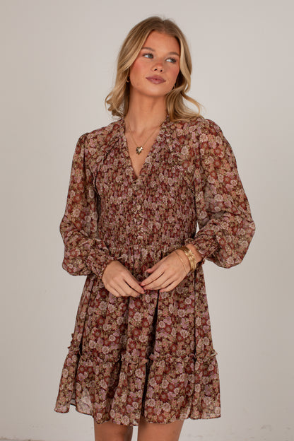 A woman modeling a long-sleeve floral mini dress with a ruffled V-neckline and gathered waist, in a brown, rust, and beige floral pattern against a plain light gray studio background.