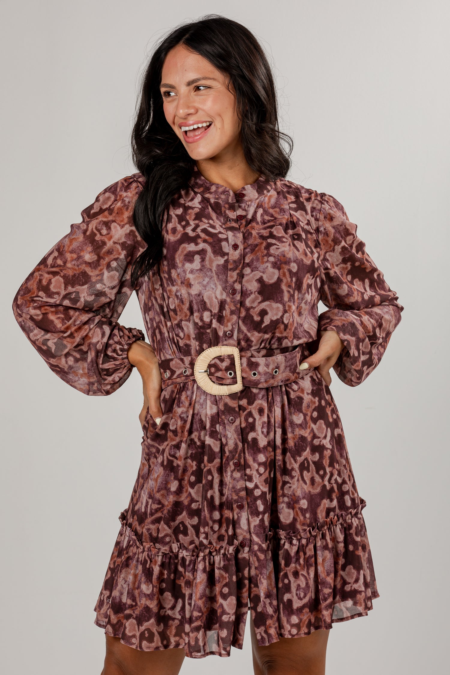 A woman modeling a long-sleeve mini dress with a brown and mauve leopard print, featuring a matching belt with a light-colored wooden buckle, set against a plain light gray background.