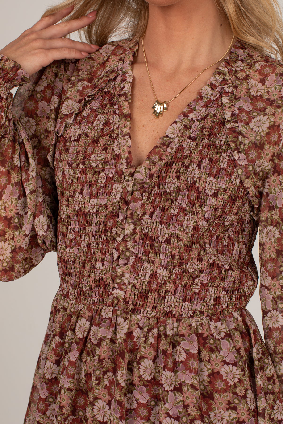 A close-up of a woman wearing a long-sleeve smocked dress with a dense floral print in muted red, pink, and beige tones, paired with a delicate gold necklace.