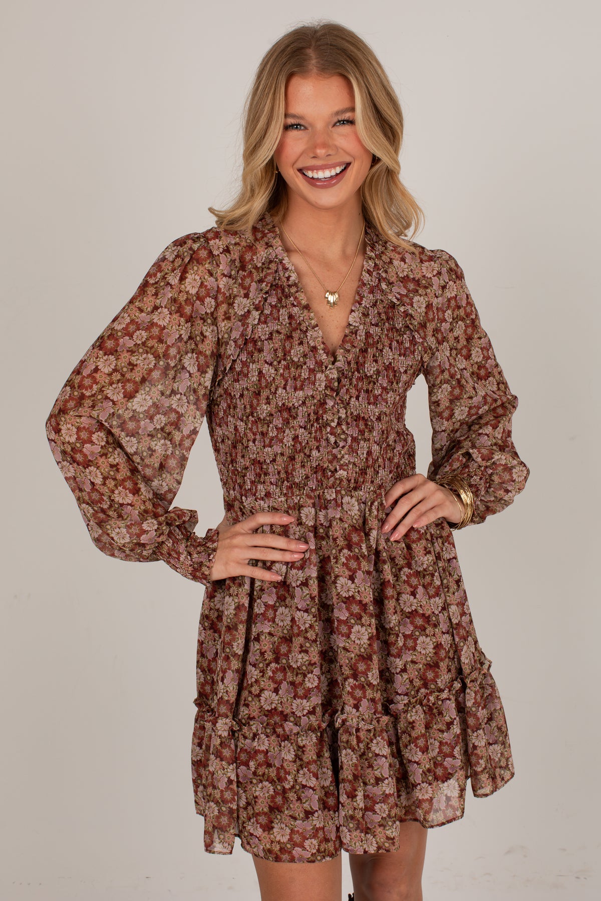 A woman modeling a long-sleeve, button-front mini dress with a brown, rust, and beige floral print against a plain light gray studio background.