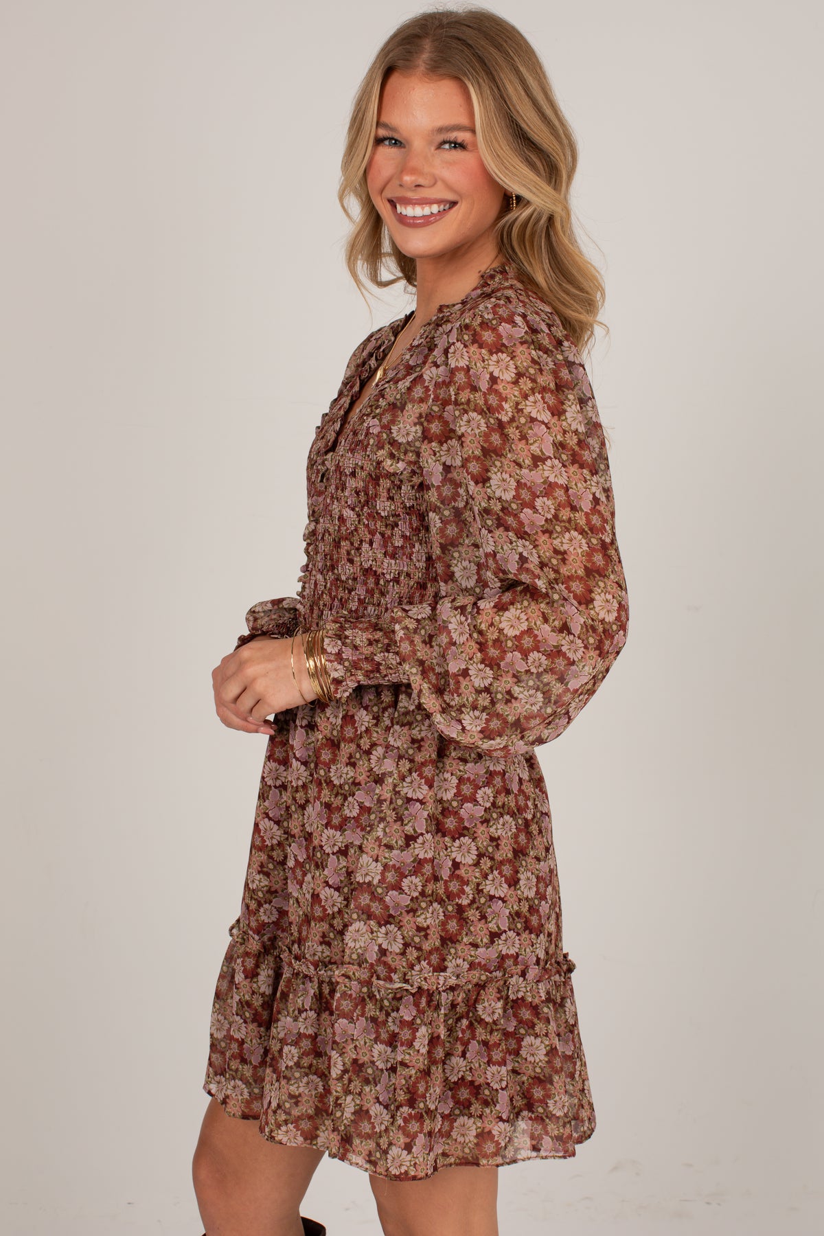 A woman modeling a long-sleeve floral mini dress with a ruffled hem, photographed against a plain light gray studio background.