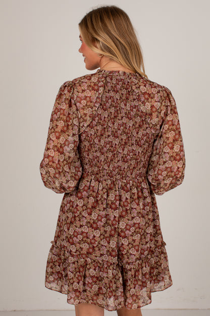 A woman viewed from the back wearing a long-sleeve smocked mini dress with a dense floral print in brown, beige, and muted red tones, set against a plain off-white background.