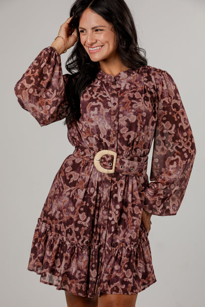 A woman modeling a long-sleeve mini dress with a brown and burgundy abstract pattern, featuring a matching waist belt with a light wood-look buckle, set against a plain light gray studio background.