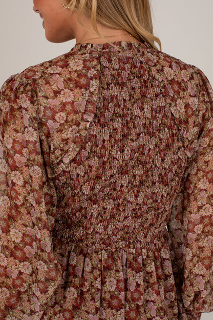 Close-up back view of a woman wearing a long-sleeve smocked dress with a dense floral pattern in dusty pink, rust red, and beige tones against a plain light background.