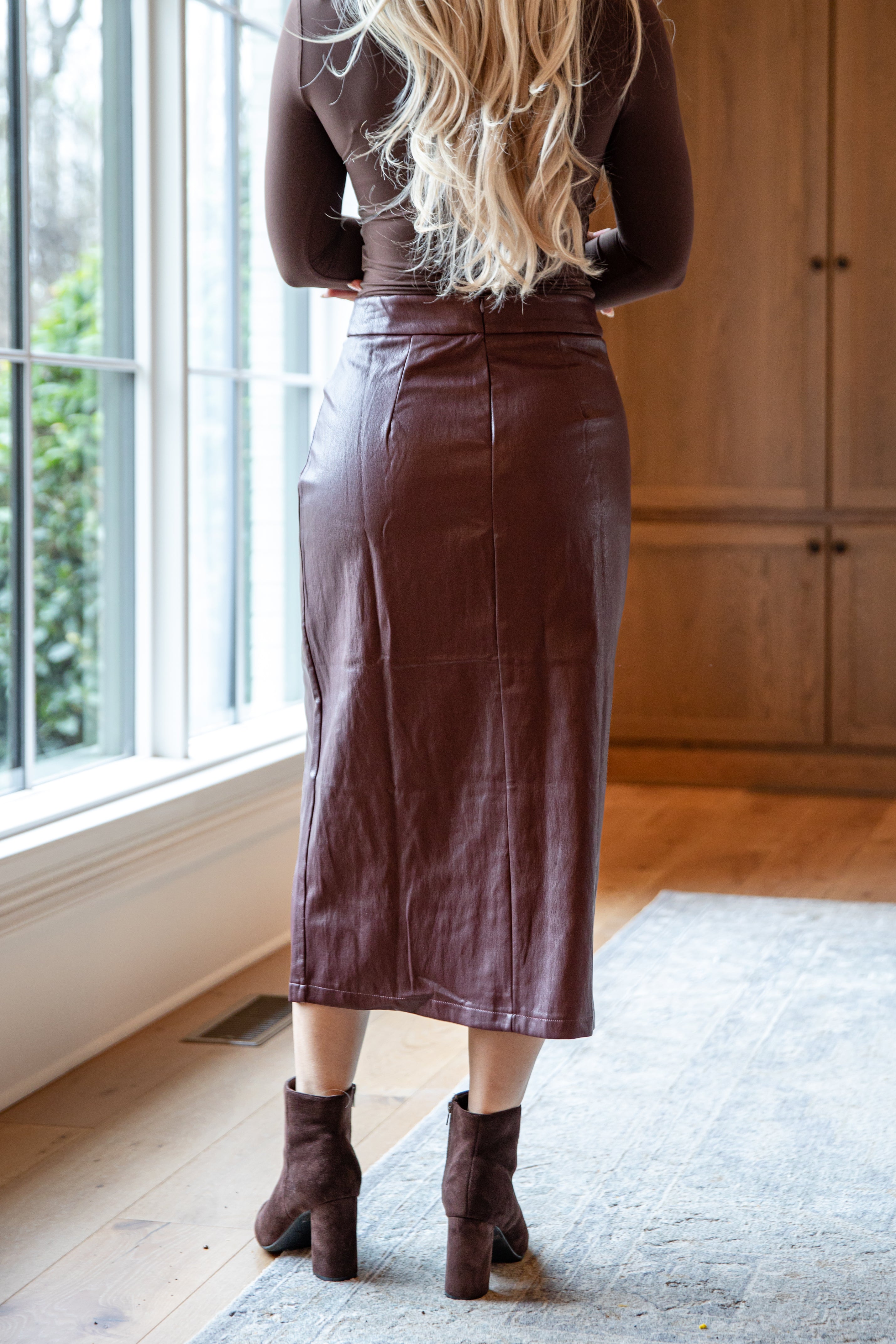 Person wearing a brown leather skirt and boots in a room with wooden flooring and a window.