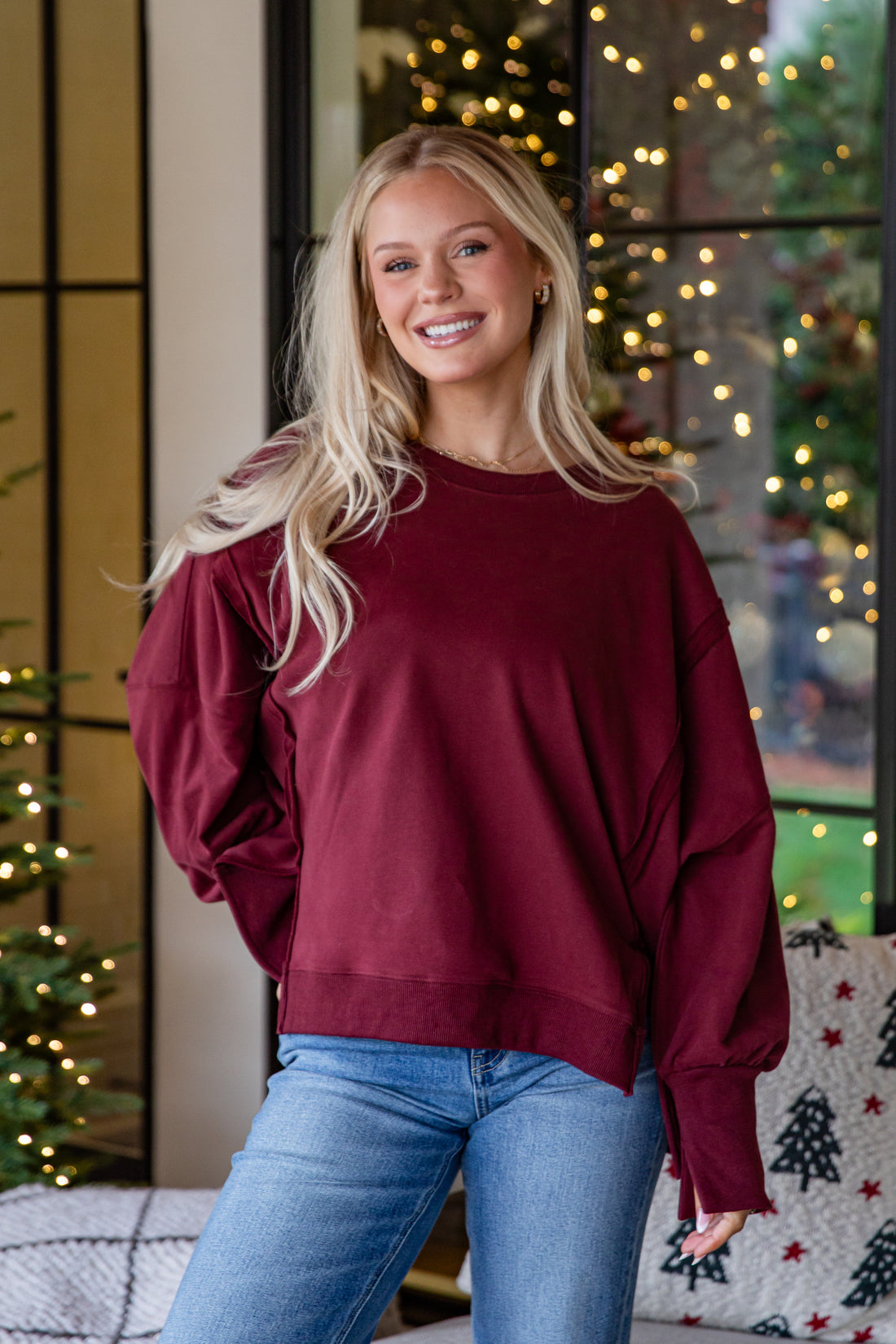 Woman wearing a burgundy sweater and blue jeans standing in front of a decorated Christmas tree.