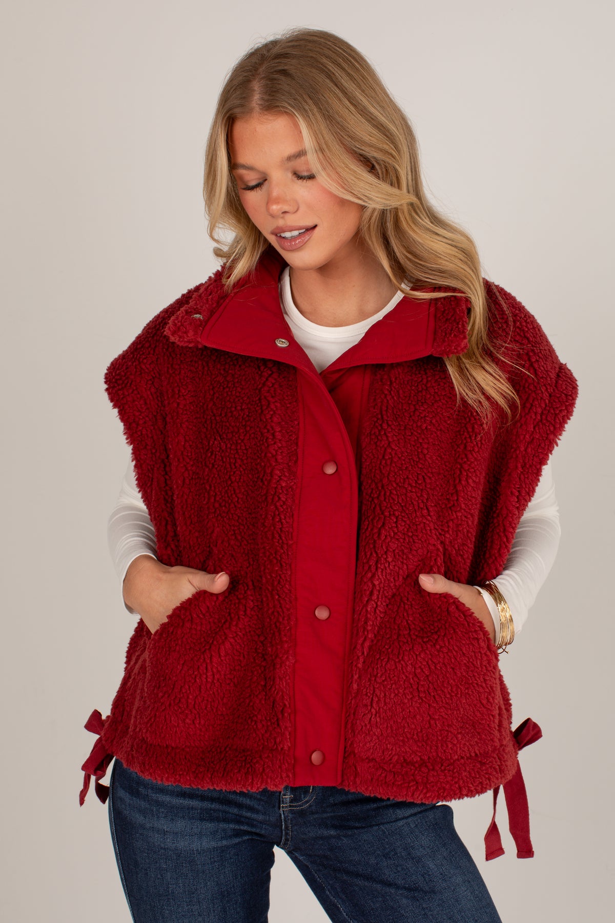 A woman wearing a red sherpa fleece button-front vest with side ties, layered over a white long-sleeve shirt and blue jeans, against a plain light gray studio background.