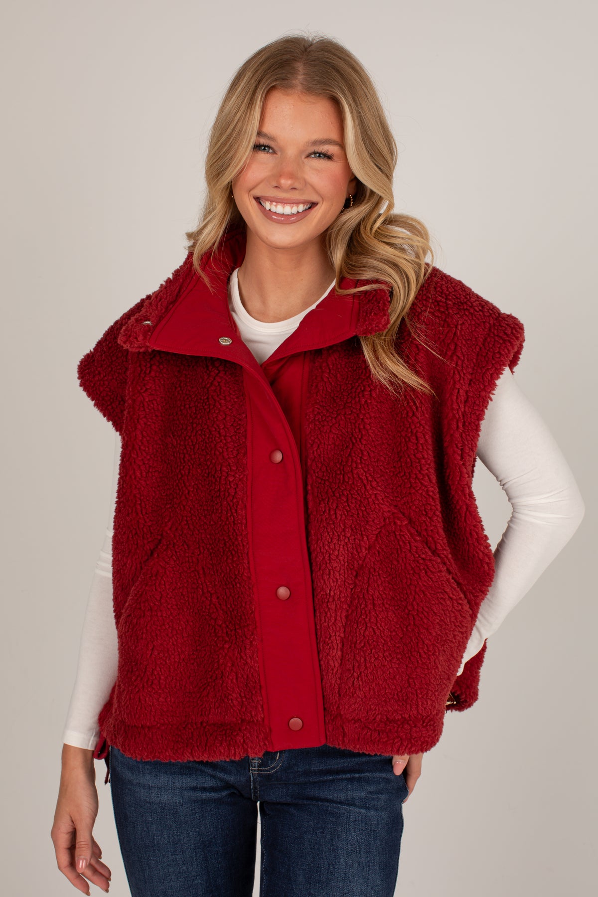 A smiling blonde woman wearing a red sherpa fleece button-front vest over a white long-sleeve top and blue jeans, photographed against a plain light gray studio background.