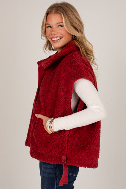 A smiling blonde woman wearing a plush red sherpa sleeveless vest over a white long-sleeve top and blue jeans, posed against a light gray studio background.