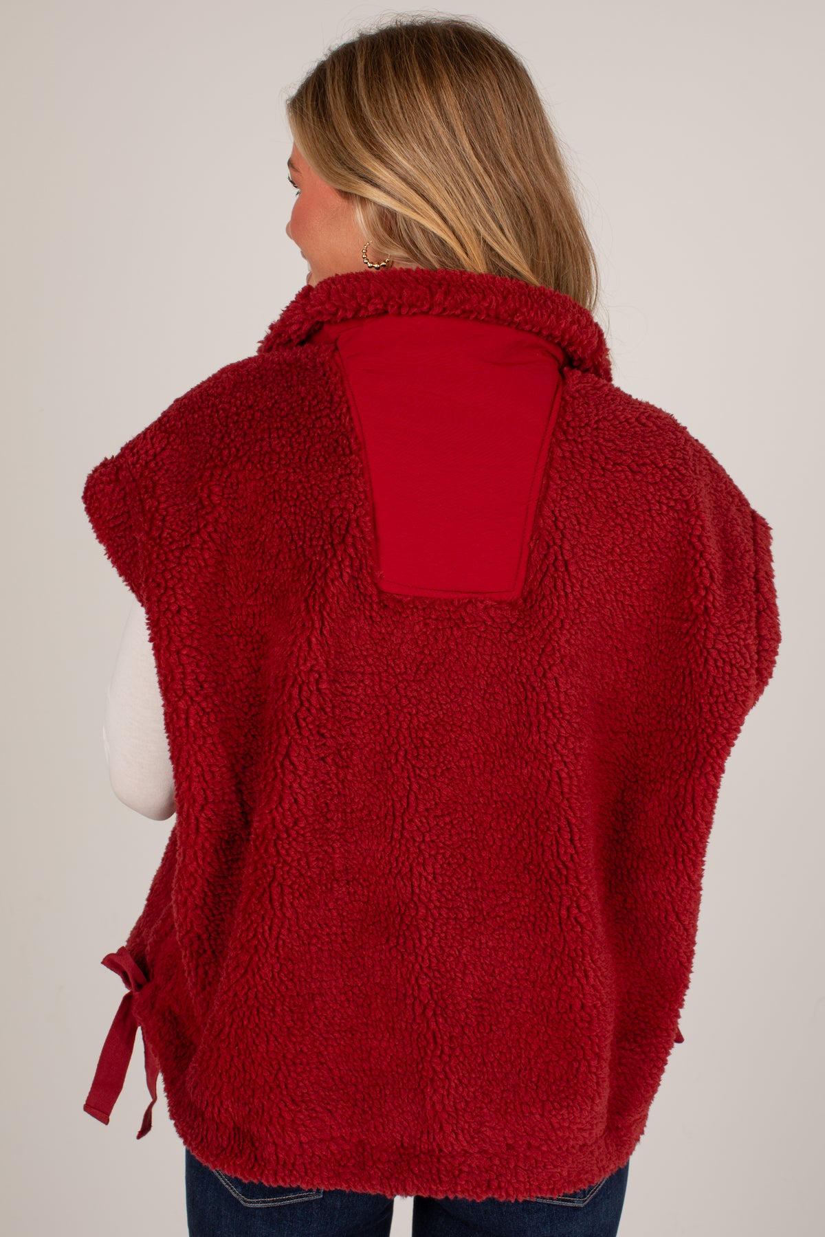 A woman shown from the back wearing a red sherpa fleece vest over a white long-sleeve top and blue jeans, against a plain light gray studio background.