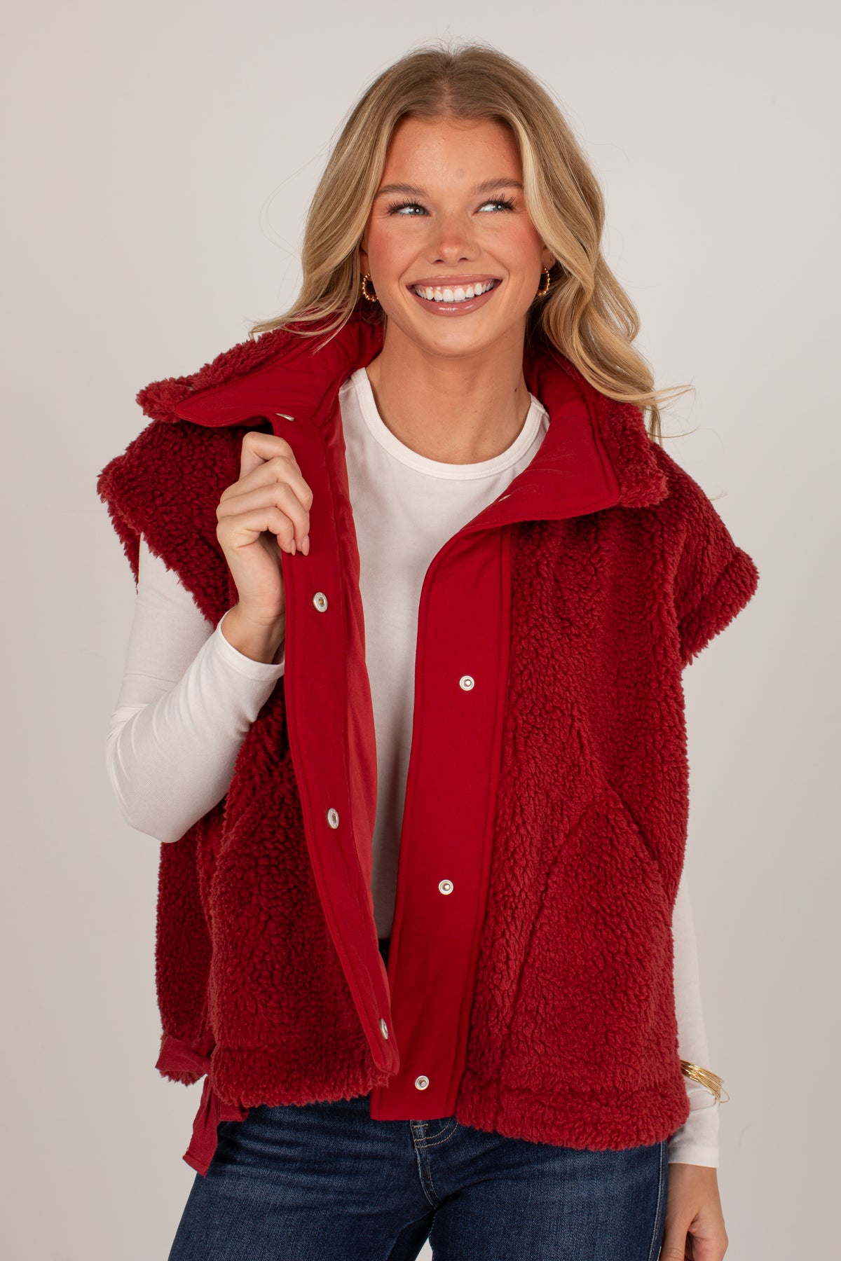 A smiling blonde woman wearing a red sherpa-lined snap-front vest over a white long-sleeve top and blue jeans, photographed against a plain light gray studio background.