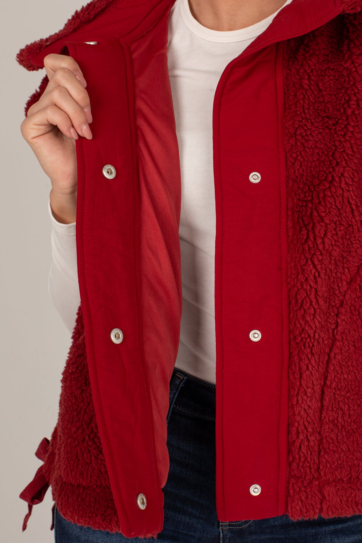 A close-up of a person wearing a red sherpa-lined jacket with a red outer panel and silver snap buttons, layered over a white shirt and blue jeans.