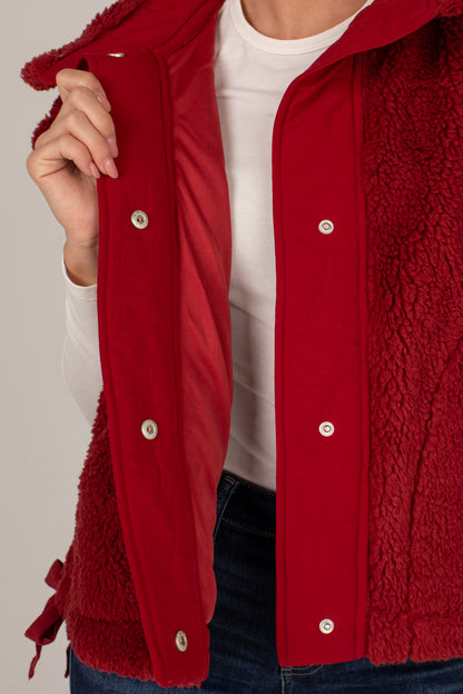 A close-up of a person wearing a red sherpa-lined jacket with a red outer panel and silver snap buttons, layered over a white shirt and blue jeans.
