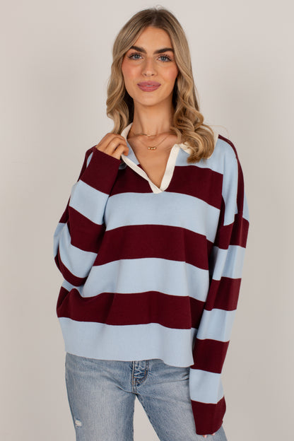 A woman wearing a light blue and burgundy striped knit sweater with a white collar detail, paired with light-wash blue jeans, against a plain light gray studio background.