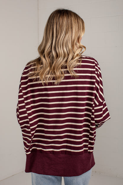 A woman viewed from behind wearing a loose-fitting short-sleeve top with horizontal burgundy and cream stripes, paired with light blue jeans, against a white brick wall.