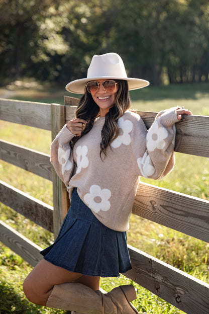 Woman wearing a floral sweater, hat, and skirt standing by a wooden fence in a field.