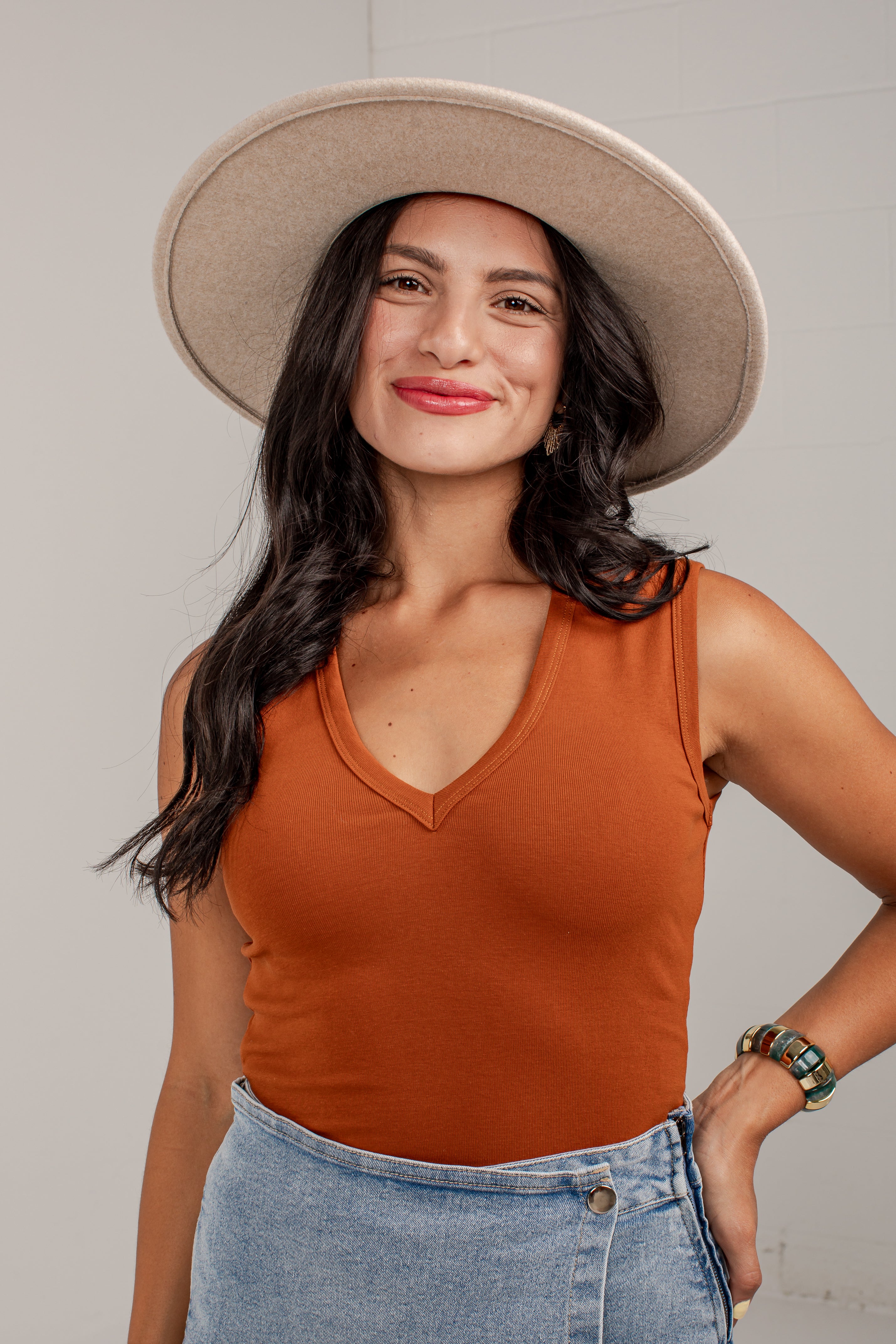 A woman wearing a ribbed sleeveless V-neck tank top in burnt orange, a wide-brimmed beige hat, and a light blue denim skirt, posing against a light gray wall.