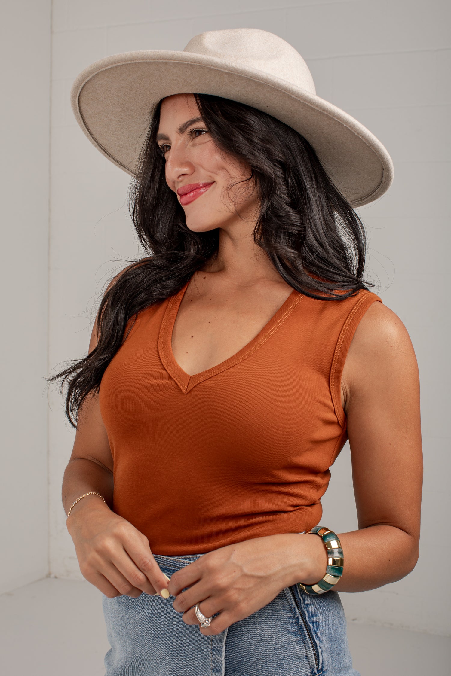 A woman wearing a rust-orange sleeveless V-neck top, a wide-brimmed cream hat, and light-wash blue jeans, standing against a light gray wall.