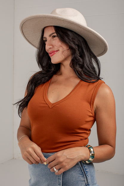 A woman wearing a rust-orange sleeveless V-neck top, a wide-brimmed cream hat, and light-wash blue jeans, standing against a light gray wall.