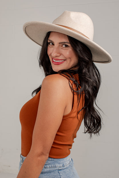 A woman wearing a wide-brimmed cream fedora hat and a rust-orange ribbed tank top, photographed against a light gray studio background.
