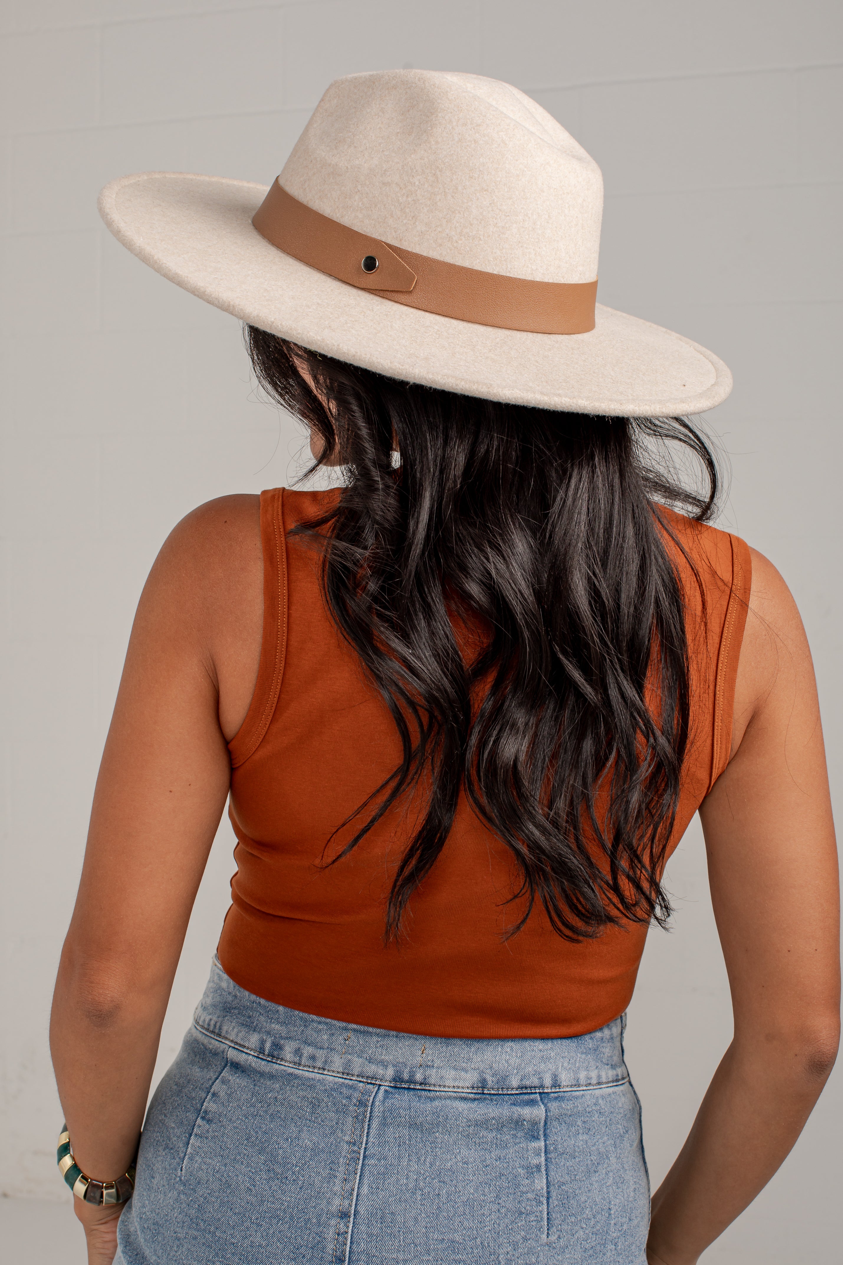 A woman shown from behind wearing a wide-brim fedora hat in cream with a tan band, a rust-orange sleeveless top, and light-wash blue jeans against a plain light gray background.