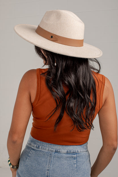 A woman shown from behind wearing a wide-brim fedora hat in cream with a tan band, a rust-orange sleeveless top, and light-wash blue jeans against a plain light gray background.