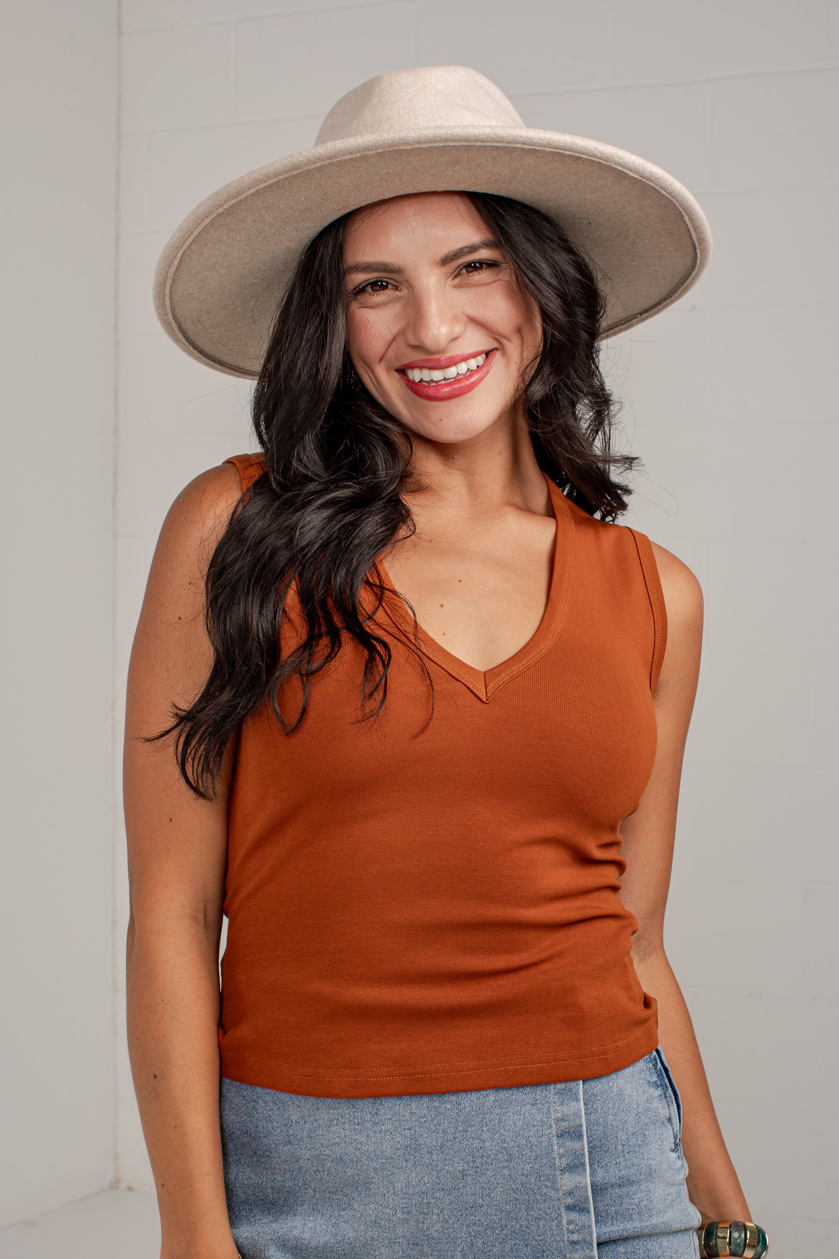 A smiling woman wearing a rust-orange ribbed sleeveless V-neck tank top, a wide-brimmed beige hat, and light-wash blue jeans, standing against a light gray wall.