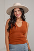 A smiling woman wearing a rust-orange ribbed sleeveless V-neck tank top, a wide-brimmed beige hat, and light-wash blue jeans, standing against a light gray wall.